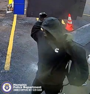 A surveillance-style image shows a person standing outdoors in a paved area marked with yellow parking lines. The person is wearing dark clothing with the hood pulled up and is lifting one arm toward the head, partially obscuring the face. A bright orange traffic cone is positioned behind the person near a large dark-colored structure. Nearby, part of a vehicle is visible. In the lower-left corner of the image, there is a Memphis Police Department seal and white text that reads “Memphis Police Department CRIMESTOPPERS 901‑528‑CASH.”