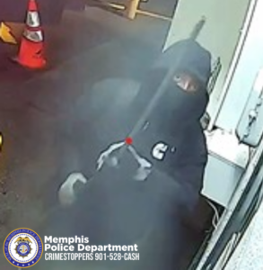 A surveillance-style image shows an individual dressed in dark clothing with a hood and a full face covering. The individual is crouched near a large machine that appears to be an ATM or kiosk. The individual is holding a long metal tool with both hands. A red dot on the tool suggests a laser pointer or targeting aid. The scene is outdoors at night, with two orange traffic cones visible in the background on a paved surface. There is smoke or dust in the air around the individual. In the bottom left corner, a round seal for the Memphis Police Department is visible beside text that reads: “Memphis Police Department CRIMESTOPPERS 901-528-CASH.”