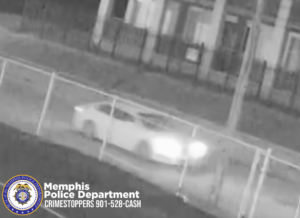 A black‑and‑white surveillance-style image shows a light-colored sedan traveling along a street at night with headlights illuminated. A metal fence runs parallel to the roadway in the foreground. In the background are several buildings with porches and columns. In the lower-left corner, the seal of the Memphis Police Department appears along with white text that reads “Memphis Police Department Crimestoppers 901‑528‑CASH.”