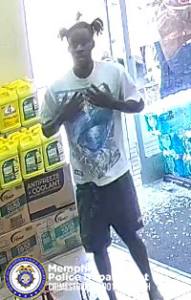 A surveillance-style image shows an individual standing near the entrance of a store. The individual is wearing a light‑colored graphic T‑shirt, dark shorts, and dark footwear. The individual has a distinctive hairstyle with multiple sections gathered into high tied portions. The person is standing with both hands held against the chest area. To the left side of the image are stacked yellow plastic containers labeled “ANTIFREEZE & COOLANT” along with other boxed products. Sunlight shines through the glass door behind the individual, and parts of promotional posters are visible through the glass. In the bottom left corner of the image, there is a circular Memphis Police Department Crime Stoppers logo.