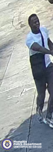 A surveillance-style image shows an individual walking outdoors on a paved surface. The individual is wearing a short‑sleeved shirt with contrasting light and dark sections, long dark pants, and athletic shoes with light-colored soles. The person has a bag or strap worn diagonally across the torso. The person’s arms are extended slightly forward as if in mid‑motion. The lower-left corner of the image contains a circular Memphis Police Department Crime Stoppers logo and contact information.