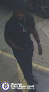 A surveillance-style image shows one individual walking on a paved surface near the edge of a yellow-painted curb. The individual is wearing dark clothing including a short-sleeved shirt, pants, and shoes, along with a large circular pendant necklace and a cap. The person is holding an object in one hand. A vehicle with large tires is partially visible in the background. In the lower corner of the image, there is a Memphis Police Department badge logo and text that reads: “Memphis Police Department CRIMESTOPPERS 901-528-CASH.”