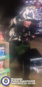 A blurry surveillance-style image shows an individual wearing a hooded garment with a bold, high-contrast pattern. The individual is bent forward near a store display containing boxed snack products. The flooring appears to be tile, and additional shelving with various packaged items is visible in the background. In the bottom corner of the image, a Memphis Police Department CrimeStoppers badge and contact information (“CRIMESTOPPERS 901-528-CASH”) are displayed.