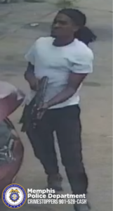 A surveillance-style image shows one individual standing outdoors on a paved surface near a vehicle. The individual is wearing a light-colored short-sleeved shirt and dark pants, with hair extending behind the head. The individual is holding a long, dark-colored object with both hands. A portion of a red vehicle is visible in the left side of the frame. In the lower corner of the image, a Memphis Police Department badge logo appears along with text that reads: “Memphis Police Department CRIMESTOPPERS 901-528-CASH.”