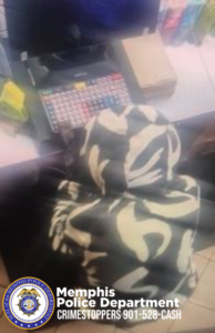 A blurry overhead surveillance-style image shows an individual wearing a hooded garment with a bold, high-contrast pattern consisting of large, curved shapes. The individual is positioned in front of a store checkout counter. On the counter is a keyboard with multiple colored keys, a monitor above it, a closed cardboard box, and several small items nearby. The flooring below appears to be tiled. In the lower corner of the image, a Memphis Police Department CrimeStoppers badge is visible, along with the text “Memphis Police Department CRIMESTOPPERS 901-528-CASH.”