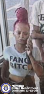An image shows an individual standing indoors near a glass door, holding a mobile phone to the side of the head. The individual has hair styled in a tall, wrapped bun that is brightly colored. The person is wearing a light‑colored shirt with large printed text that reads “FANG BITCH.” Another person, whose torso is partially visible, stands nearby wearing a shirt with dark printed letters. At the bottom of the image, there is a Memphis Police Department emblem and text that reads “Memphis Police Department CRIMESTOPPERS 901-528-CASH.”