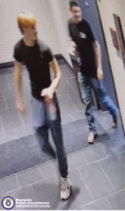 The image appears to be a security‑camera still taken in an indoor hallway with tiled flooring. Two individuals are walking through the hallway. The individual in the foreground is wearing a short‑sleeved dark shirt, long pants, and light‑colored shoes. This person has short hair and is holding an object in one hand. The second individual is behind and slightly to the right, also wearing a dark short‑sleeved shirt, long pants, and light‑colored shoes. The second individual is holding an item in one hand while walking. A Memphis Police Department CrimeStoppers logo and the text "Memphis Police Department CRIMESTOPPERS 901‑528‑CASH" are displayed in the lower left corner.