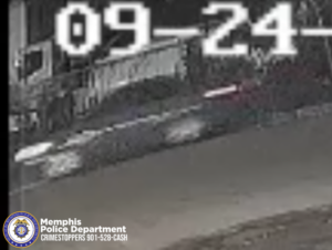 A low‑resolution black‑and‑white surveillance image shows a dark-colored car driving on a roadway at night. The vehicle appears slightly blurry due to motion. Large white timestamp text is displayed at the top of the image, reading “09‑24”. In the lower-left corner, there is a Memphis Police Department badge and text that says “Memphis Police Department CRIMESTOPPERS 901‑528‑CASH.” The background includes indistinct shapes of buildings or parked vehicles.