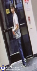 A security‑camera image shows an individual standing in front of a glass door inside a building. The individual is wearing a short‑sleeved shirt with a patterned design, long pants, and athletic shoes. The individual is holding an object in one hand while turning slightly toward the camera. On the wall beside the door, there are posted signs, including a red sign above a smaller device or panel. The floor is tiled. In the lower left corner of the image, there is a Memphis Police Department CrimeStoppers logo and the text “Memphis Police Department CRIMESTOPPERS 901‑528‑CASH.”