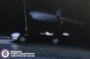 A dark, low‑resolution nighttime surveillance image shows a vehicle moving along a roadway. Only portions of the vehicle are visible due to motion blur and limited lighting; faint reflections of the headlights and wheels can be seen near the bottom of the frame. The background is mostly black, with a vertical light-colored pole or post visible on the left side. In the bottom-left corner, the Memphis Police Department badge appears beside text that reads “Memphis Police Department CRIMESTOPPERS 901‑528‑CASH.”