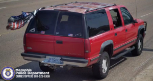 A red SUV is traveling on a multi‑lane road. The vehicle has dark‑tinted windows and a roof rack. The rear license plate is visible, along with a trailer hitch attachment underneath the bumper. Several decals are on the back window, including small flag designs and other stickers. A large flag with a striped pattern is mounted on the left side of the rear of the vehicle and is extended outward by the wind. The SUV has large tires and dark trim along the wheel wells. In the lower left corner of the image, there is a Memphis Police Department CrimeStoppers logo and the text “Memphis Police Department CRIMESTOPPERS 901‑528‑CASH.”