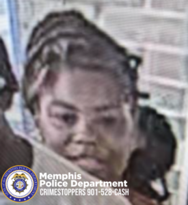 An image shows an individual positioned near what appears to be a brick wall and a window or glass door. The individual has hair styled with multiple sections or braids visible and is wearing a light‑colored top. Another person is partially visible on the left edge of the frame. The background includes brick detailing and a reflective glass surface. At the bottom of the image, a Memphis Police Department emblem is shown along with text that reads “Memphis Police Department CRIMESTOPPERS 901‑528‑CASH.”