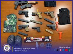 A tabletop display of confiscated items arranged neatly on a wooden surface. The items include multiple firearms of various types, including several handguns, extended magazines, and one larger rifle-style weapon at the top. Yellow safety ties are threaded through the barrels of several guns. Also displayed are ammunition boxes, loose rounds, three smartphones, two ski masks—one camouflage-patterned and one black with a white swoosh logo—an orange emergency seatbelt cutter, a pair of black slip‑on shoes resembling Crocs, and assorted accessories including a watch and small patches. The bottom left corner contains the Memphis Police Department seal, and the bottom center reads “Memphis Police Department — Established in 1827.” Social media icons and the text “joinmpd.com” appear in the lower right.