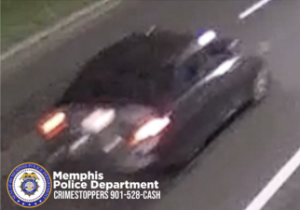 A surveillance image shows a dark-colored sedan driving on a roadway at night. The vehicle is captured from behind at an angle, with illuminated tail lights and a bright reflection on the trunk area. The road markings include two white lane lines running parallel along the right side of the frame. A grassy area is visible in the upper left corner. The bottom left of the image displays the Memphis Police Department CrimeStoppers logo and the text “Memphis Police Department CRIMESTOPPERS 901-528-CASH.