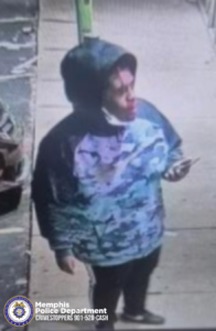 A surveillance-style image shows an individual standing on a sidewalk near a building entrance. The individual is wearing a hooded jacket with a blue and purple pattern, dark pants, and shoes. The individual is holding a phone in one hand and a face covering rests below the chin. The background shows part of a parked vehicle and a storefront. In the lower-left corner, there is a Memphis Police Department seal along with the text “Memphis Police Department CRIMESTOPPERS 901‑528‑CASH.”