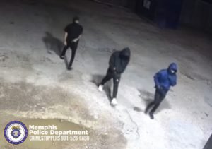 A nighttime surveillance-camera image shows three unidentified individuals walking across a concrete lot. All three individuals have their faces covered. The individual on the left is wearing dark clothing. The individual in the center is wearing a dark hooded top and light-colored shoes. The individual on the right is wearing a blue hooded top and dark pants. The ground is rough with cracks and a puddle in the foreground. In the lower-left corner is a Memphis Police Department CrimeStoppers logo and text that reads “Memphis Police Department CRIMESTOPPERS 901‑528‑CASH.”