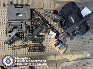 A collection of firearms, firearm components, magazines, ammunition, and gear is displayed on an outdoor perforated metal table. Items include several handguns, two rifle-style firearms, multiple magazines, ammunition boxes, and a black soft-sided gear bag labeled “Kobalt.” A hard foam-lined pistol case is open, showing a handgun and accessories inside. Loose ammunition and loaded magazines are arranged near the front of the display. In the lower-left corner, the Memphis Police Department seal is visible along with the text: “Memphis Police Department — Crimestoppers 901-528-CASH.”