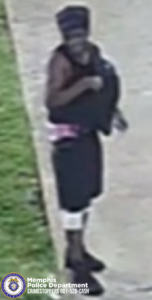 A surveillance-style image showing a person standing on a sidewalk next to a grassy area. The person is wearing a tall head covering with horizontal stripes, a sleeveless top, dark shorts, light-colored fabric wrapped around the lower legs, and dark shoes. The person is holding or adjusting an item near the upper body. The image includes a Memphis Police Department CrimeStoppers logo and the text: “Memphis Police Department CRIMESTOPPERS 901-528-CASH.”