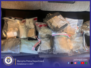 The image shows numerous sealed evidence bags arranged across a dark surface. Each bag contains different materials: some appear to hold white or off‑white powder, some contain solid block-like substances, others contain granulated or pellet-like material, and several bags contain multiple smaller interior bags. Many of the evidence bags have printed labels attached, including forms labeled “Chain of Custody,” though the handwritten fields are not readable. Some bags are resealable plastic, while others appear heat‑sealed or wrapped in reflective material. The items are laid out in rows, taking up most of the frame. The background includes part of a light gray upholstered surface at the top of the image. Along the bottom of the image is a dark purple banner displaying the Memphis Police Department seal on the left, accompanied by the text: “Memphis Police Department – Established in 1827.” On the right side of the banner is the text “joinmpd.com” and a row of small social media icons.