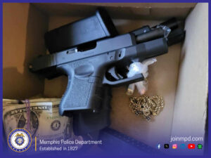 “Inside a cardboard box, there is a black semi‑automatic handgun with the slide slightly pulled back. Nearby are bundled U.S. dollar bills, a small gold-colored chain, and several small plastic baggies containing unidentified items. The lower-left corner features the Memphis Police Department seal with the text ‘Memphis Police Department, Established in 1827.’ The lower-right corner includes the website ‘joinmpd.com’ and various social media icons.”