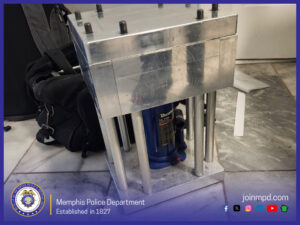 “An aluminum mechanical press-like device stands on a tile floor. The structure has a rectangular metal top plate secured with several bolts and supported by multiple vertical metal rods. Between the rods is a blue hydraulic bottle jack labeled ‘Duralast 12‑Ton’. A black bag sits partially visible behind the device. At the bottom left is the Memphis Police Department seal with the text ‘Memphis Police Department, Established in 1827,’ and at the bottom right is the website ‘joinmpd.com’ with social media icons.”