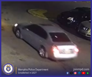 A surveillance-style photograph showing the rear three-quarter view of a silver or light-gray four-door sedan driving along an asphalt street at night. The car's headlights and rear tail/license plate lights are illuminated, and other vehicles are parked along the curb in the upper-right background. Across the bottom banner is the Memphis Police Department seal with the text "Memphis Police Department – Established in 1827", alongside the website "joinmpd.com" and social media icons.