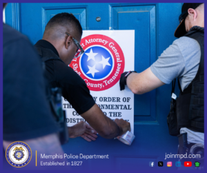 Two law‑enforcement personnel are taping a printed notice to a blue door. The notice features a circular emblem with three stars and the text “Attorney General” and “Shelby County, Tennessee.” One individual holds the lower part of the notice while applying tape, and another secures the upper edge. Both individuals are wearing tactical vests and equipment. The bottom of the image includes the Memphis Police Department seal, the text “Memphis Police Department – Established in 1827,” the website “joinmpd.com,” and several social media icons.