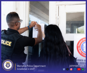 A law‑enforcement officer is taping a printed notice to a glass door. Another individual with long dark hair stands nearby, facing the door. Part of an official sign with the circular Shelby County District Attorney General emblem is visible on the right side of the image. The lower portion includes the Memphis Police Department seal, the text “Memphis Police Department — Established in 1827,” the website “joinmpd.com,” and several social media icons.