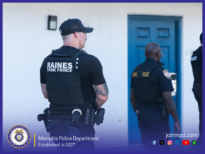 Two uniformed law‑enforcement personnel stand outside a building with a blue door. One individual in the foreground wears a vest labeled “RAINES TASK FORCE,” along with tactical gear and a cap. Another individual nearby wears a vest labeled “MPD POLICE.” The scene includes additional officers partially visible. At the bottom of the image are logos and text for the Memphis Police Department, including the department seal, the statement “Established in 1827,” and the website “joinmpd.com.” Icons for several social media platforms also appear.