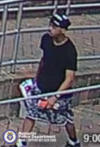  A surveillance-style image shows a person walking outdoors along a metal railing on a paved surface. The person is wearing a dark short-sleeved shirt, patterned knee-length shorts, and a hat with light-colored markings. The person is holding several items, including a red object and what appears to be packaging with bright colors. In the bottom left corner, there is a Memphis Police Department badge graphic alongside the text “Memphis Police Department CRIMESTOPPERS 901-528-CASH.” A timestamp reading “9:00” appears in the bottom right corner.