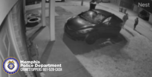 A black‑and‑white security camera image shows a parked vehicle in a driveway at night. Two individuals are walking on the right side of the frame near the vehicle. The scene appears to be recorded by a Nest camera. In the bottom-left corner, there is a Memphis Police Department badge and text that reads: “Memphis Police Department — Crimestoppers 901‑528‑CASH.” The surrounding area includes a building wall on the left, a support post, a driveway, and what appears to be a residential neighborhood in the background.