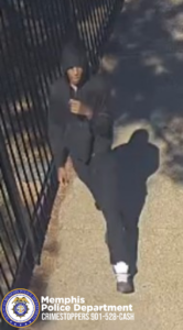 Surveillance image released by the Memphis Police Department showing an individual walking along a paved walkway beside a black metal fence, wearing a dark hooded sweatshirt with the hood up, dark pants, and white athletic shoes. The bottom left features the Memphis Police Department seal with text reading "Memphis Police Department CRIMESTOPPERS 901-528-CASH".